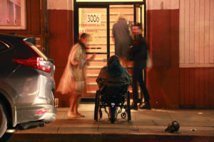 Jaklin sits in her wheelchair, looking away from the camera, and looking into a building with an open glass door, with a flight of stairs inside. It is nighttime, and there are two people who are blurry in the frame that are talking to Jaklin or looking on at her sitting there.