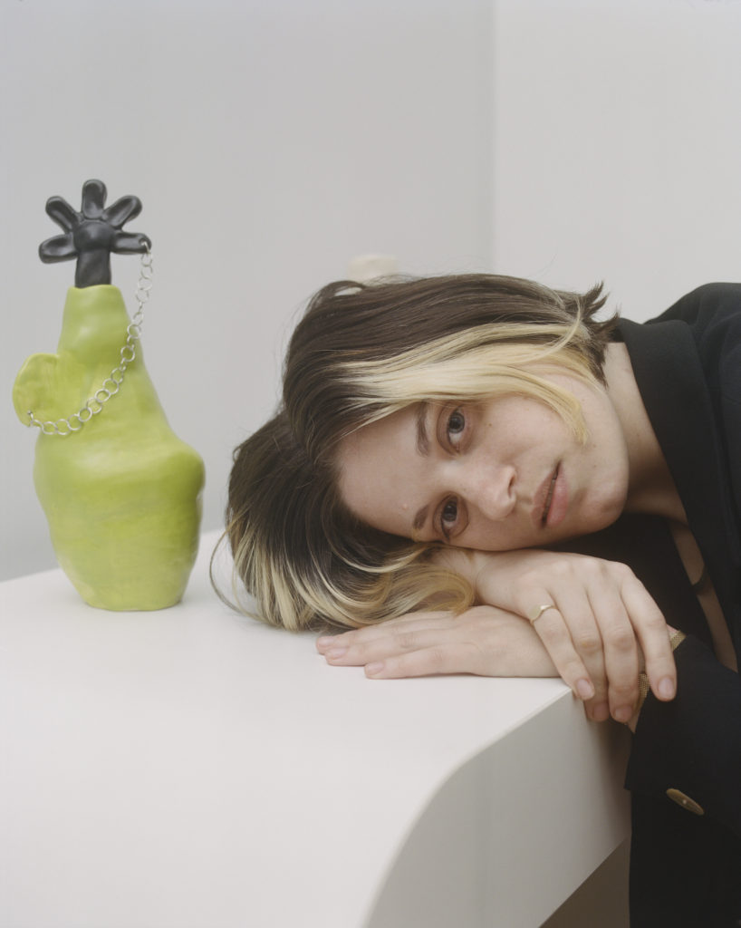 Hayley rests her head on her arms atop a white table. Next to her is one of her ceramic vessels entitled “i often mis-take my dreams for memories,” which is a lumpy bright lime green vessel with an ear-like handle. The handle is pierced and has a chain through it, which connects to a black ceramic flower petal that is atop it.