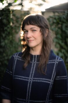 White woman with brown hair and bangs, and a blue and grey sweater.