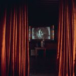 Orange-red curtains framing the screen for a black and white movie that depicts a woman on the screen, the projector on a table in front of it.