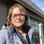 White woman with blond hair, brown glasses, and light blue jacket