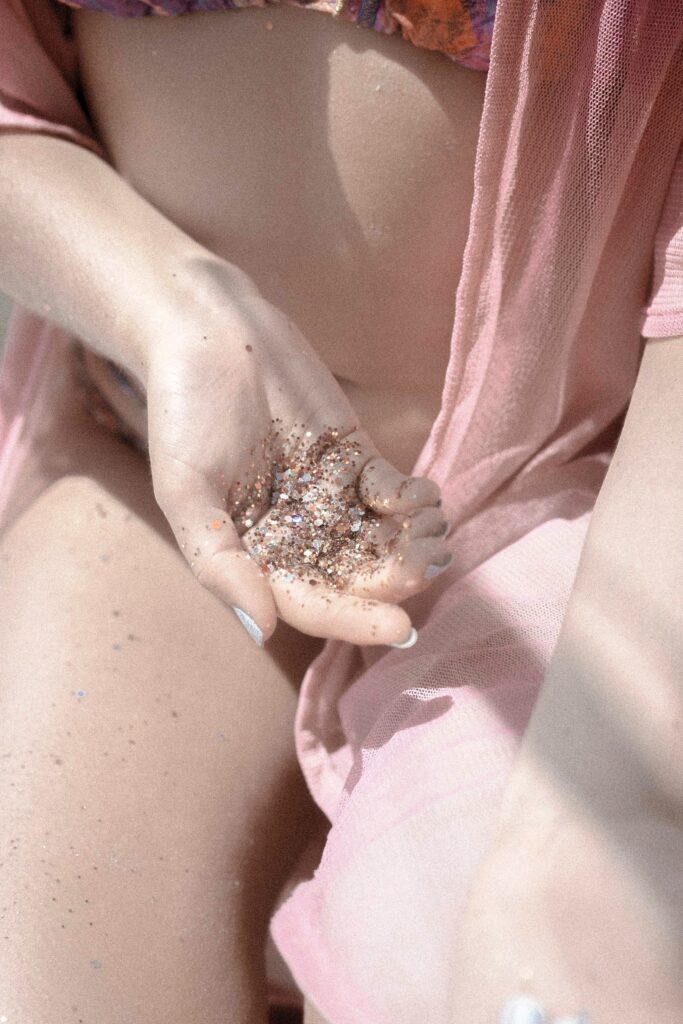 Woman holds pile of of silver and gold glitter in her hands with fingernails painted white, wearing an orange and pink patterned bikini and light pink sheer cover up