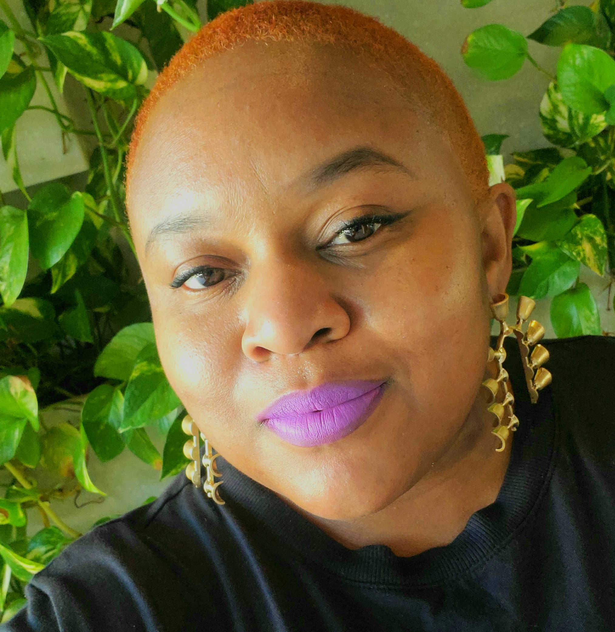 Photo of a black woman with purple lipstick, hanging gold earrings, and orange buzzed hair.