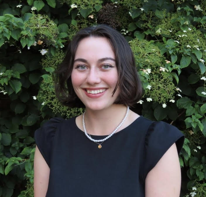 White woman with dark brown hair and black top, light green eyes.