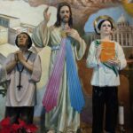 Photo of three figurines, the middle figure appears to be Jesus, wearing a beige tunic and with a gold circle emitting pink and blue rays downwards. There are white clouds, brown mountains in the background. The figure on the left has hands clasped, a white top, and a black necklace with a cross on it. The figure on the right is also wearing a white top with a green fern-like branch across it, a gold halo around his head, and an orange book with the words "Doctrina Cristiana"