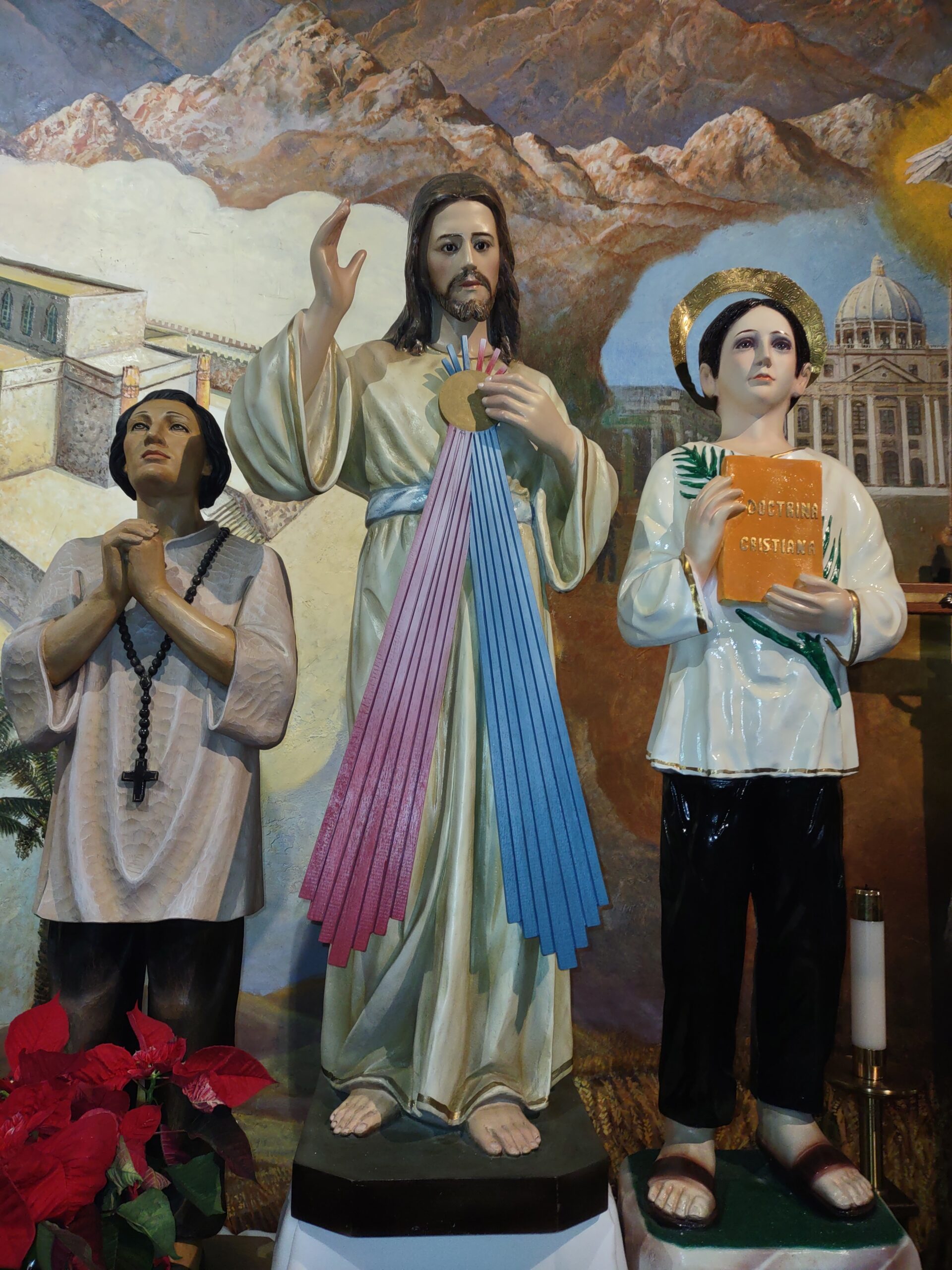 Photo of three figurines, the middle figure appears to be Jesus, wearing a beige tunic and with a gold circle emitting pink and blue rays downwards. There are white clouds, brown mountains in the background. The figure on the left has hands clasped, a white top, and a black necklace with a cross on it. The figure on the right is also wearing a white top with a green fern-like branch across it, a gold halo around his head, and an orange book with the words "Doctrina Cristiana"