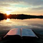 Image of a book on a brown wooden table by the lake with the sunset and trees by the horizon.
