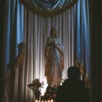 A person kneeling in prayer before a statue of the Virgin Mary in a dimly lit space, with several lit votive candles on a table in the foreground.