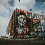 A large, colorful mural of a smiling Anne Frank with multicolored stripes across her face covers the side of a tall brick industrial building. The words “Let me be myself” appear above the portrait. The building is surrounded by graffiti, with a few cars parked along the street under a partly cloudy sky.