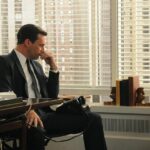 Don Draper from AMC's Mad Men in a dark suit sits pensively at his office desk, resting his chin on his hand as he looks down. Behind him, large windows with venetian blinds reveal tall city buildings in soft daylight.