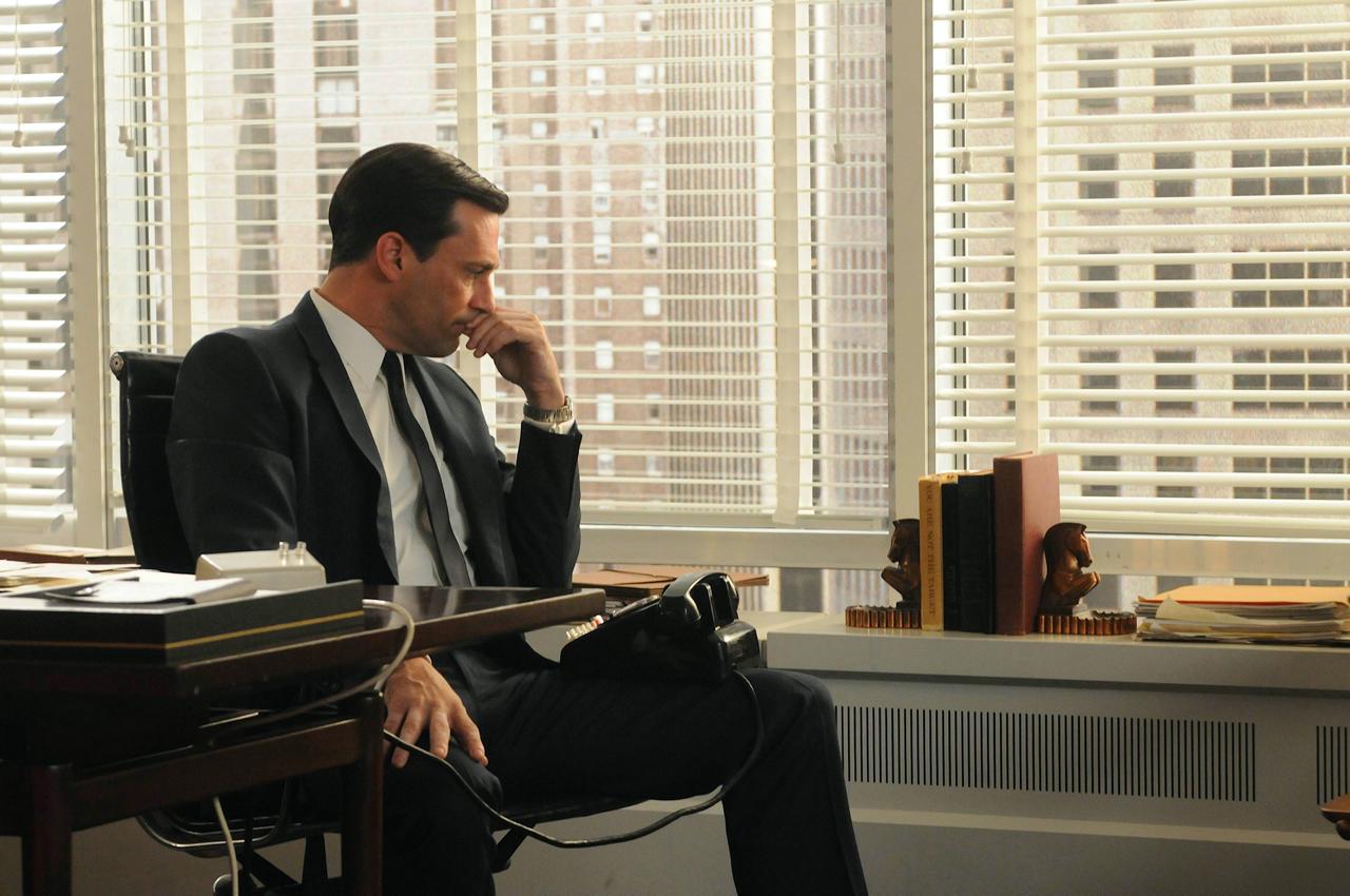 Don Draper from AMC's Mad Men in a dark suit sits pensively at his office desk, resting his chin on his hand as he looks down. Behind him, large windows with venetian blinds reveal tall city buildings in soft daylight.