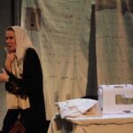 A woman wearing a traditional embroidered dress and a light headscarf performs on stage beside a white sewing machine and folded cloth. Behind her hangs a backdrop covered in handwritten Arabic text.