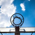 Upward view of an old, weathered basketball hoop without a net against a bright blue sky with scattered white clouds.