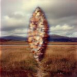 A translucent, glitching triangular form composed of distorted digital pixels stands vertically in a rural moorland landscape with golden grasses, a water body, and distant mountains under a cloudy sky.