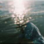 Person with long hair wearing a blue sweatshirt sits by the water with their head down, sunlight creating bokeh effects across the image