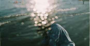 Person with long hair wearing a blue sweatshirt sits by the water with their head down, sunlight creating bokeh effects across the image