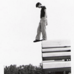 Black and white photograph of a man with short hair in pants, a T-shirt, and flip-flop sandals standing at the top of bleachers on the very edge, looking down, with one foot in the air as if stepping onto a tightrope.
