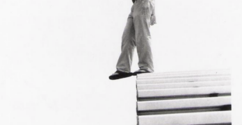 Black and white photograph of a man with short hair in pants, a T-shirt, and flip-flop sandals standing at the top of bleachers on the very edge, looking down, with one foot in the air as if stepping onto a tightrope.