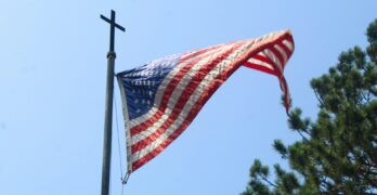 An American flag on a pole topped with a Christian cross, viewed from below