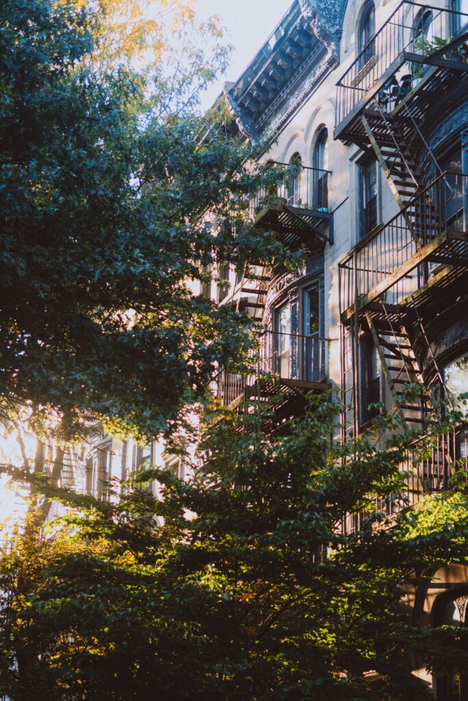 A photo of the outside of an apartment building with a fire escape, light filtering in through the trees surrounding it.