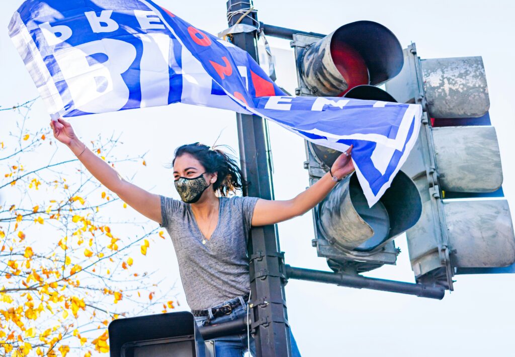A woman wearing a face mask stands on a street light and holds a blue "Biden for President 2020" banner that waves in the air.